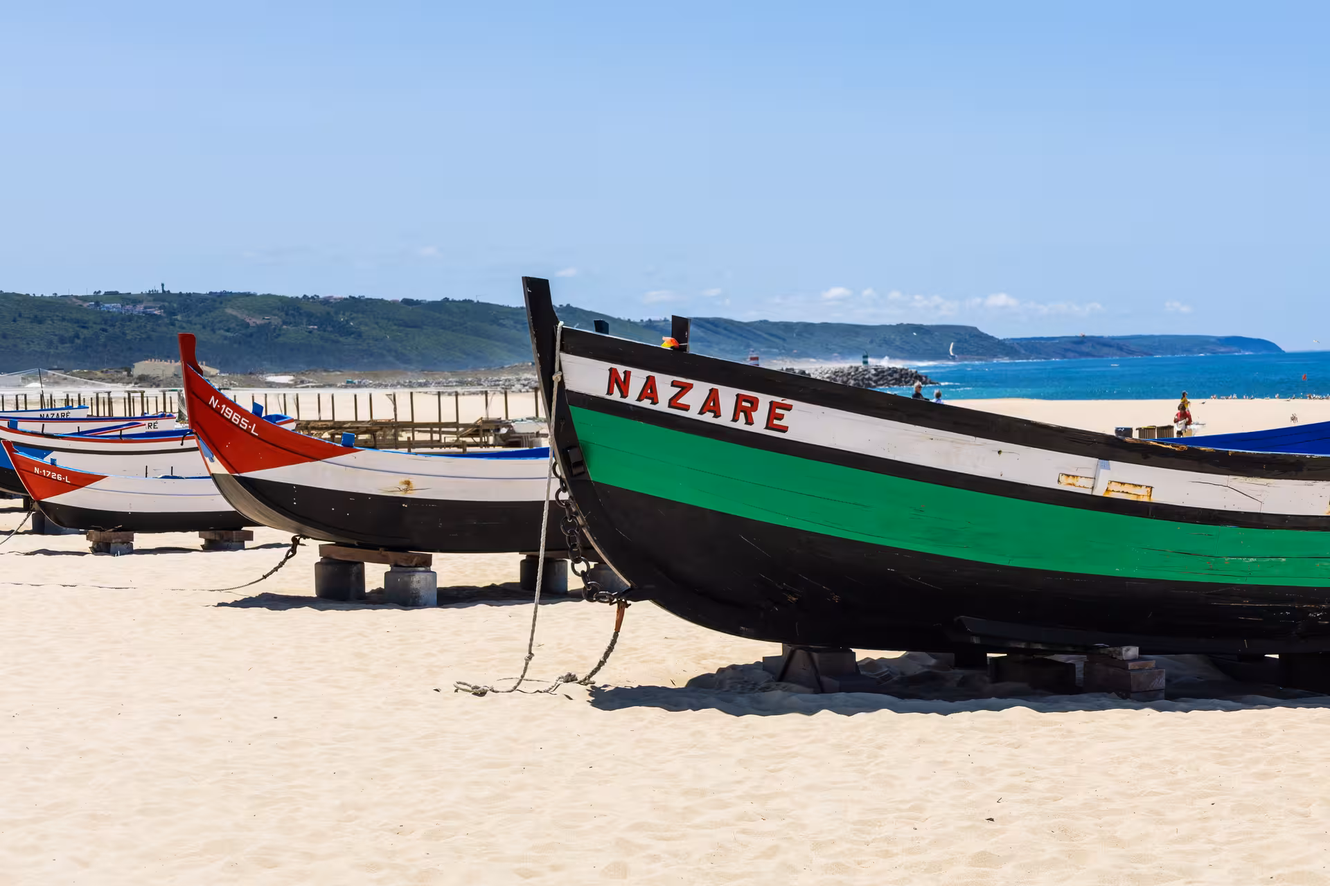 Traditional colorful fishing boats resting on the sandy shores of Nazaré, with the Atlantic Ocean and hills in the background.