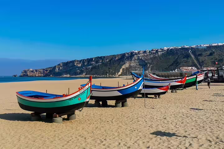 Colorful traditional fishing boats on the sandy beach of Nazaré, Portugal, with scenic cliffs in the background, perfect for a Lisbon day tour.