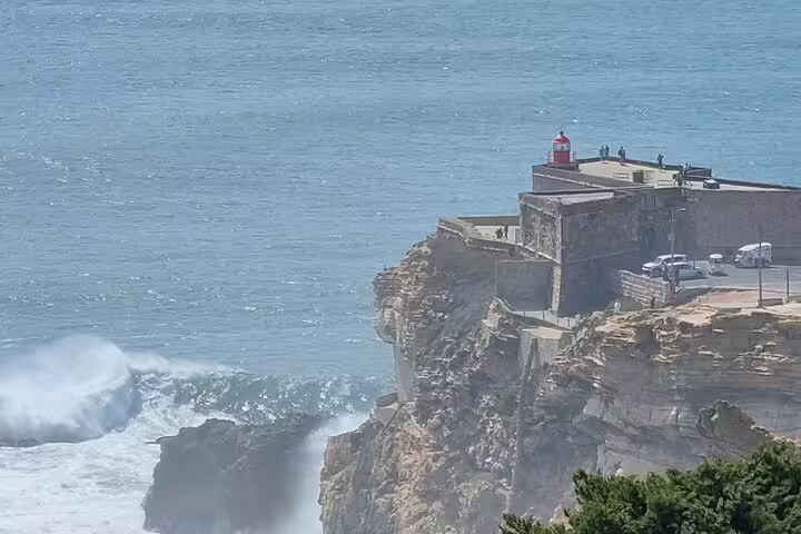 Scenic view of the iconic lighthouse on the cliffs of Nazaré, Portugal, with waves crashing against the rocky coast.