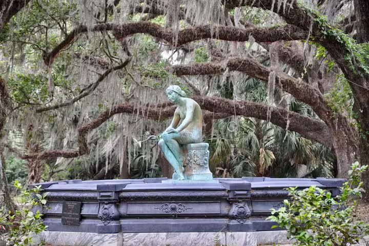 Bronze seated figure statue atop a granite tomb surrounded by Spanish moss-draped oak trees in a city park