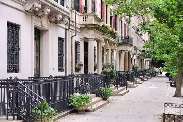 Historic brownstone townhouses with ornate ironwork railings lining a tree-shaded New York City sidewalk