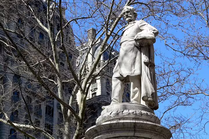 White marble statue on pedestal in New York City park with bare trees and historic building in background