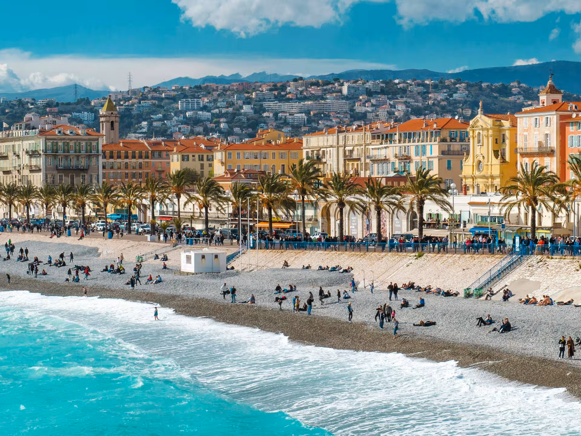 Panoramic view of Nice beach promenade with turquoise Mediterranean waters, palm trees, and colorful French Riviera buildings