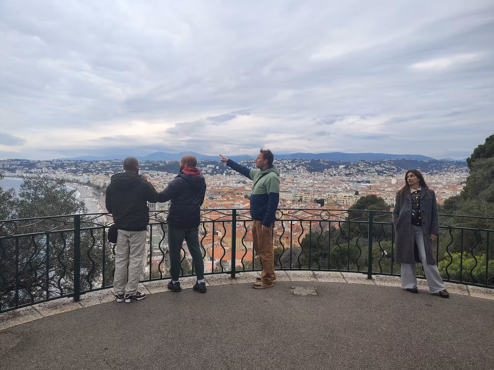 Tourists at Castle Hill viewpoint overlooking Nice city rooftops, coastline, and mountains on a cloudy day
