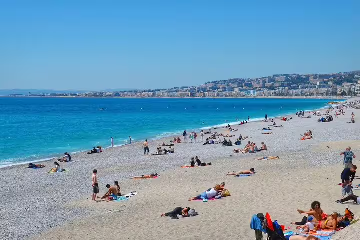 Crowded pebble beach in Nice, France with turquoise Mediterranean Sea and city skyline in background