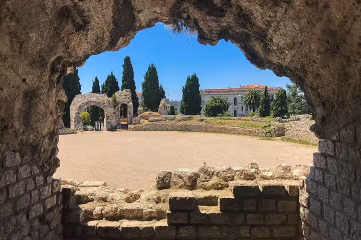 Ancient Roman ruins arena viewed through stone archway with cypress trees and historic building in Nice