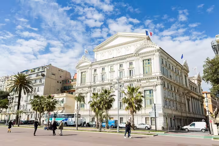 Ornate white Opéra de Nice building with French flag, palm trees, and pedestrians on sunny day