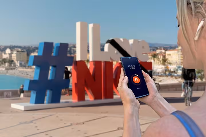 Woman holding smartphone with audio guide app near I Love Nice landmark sign on the promenade