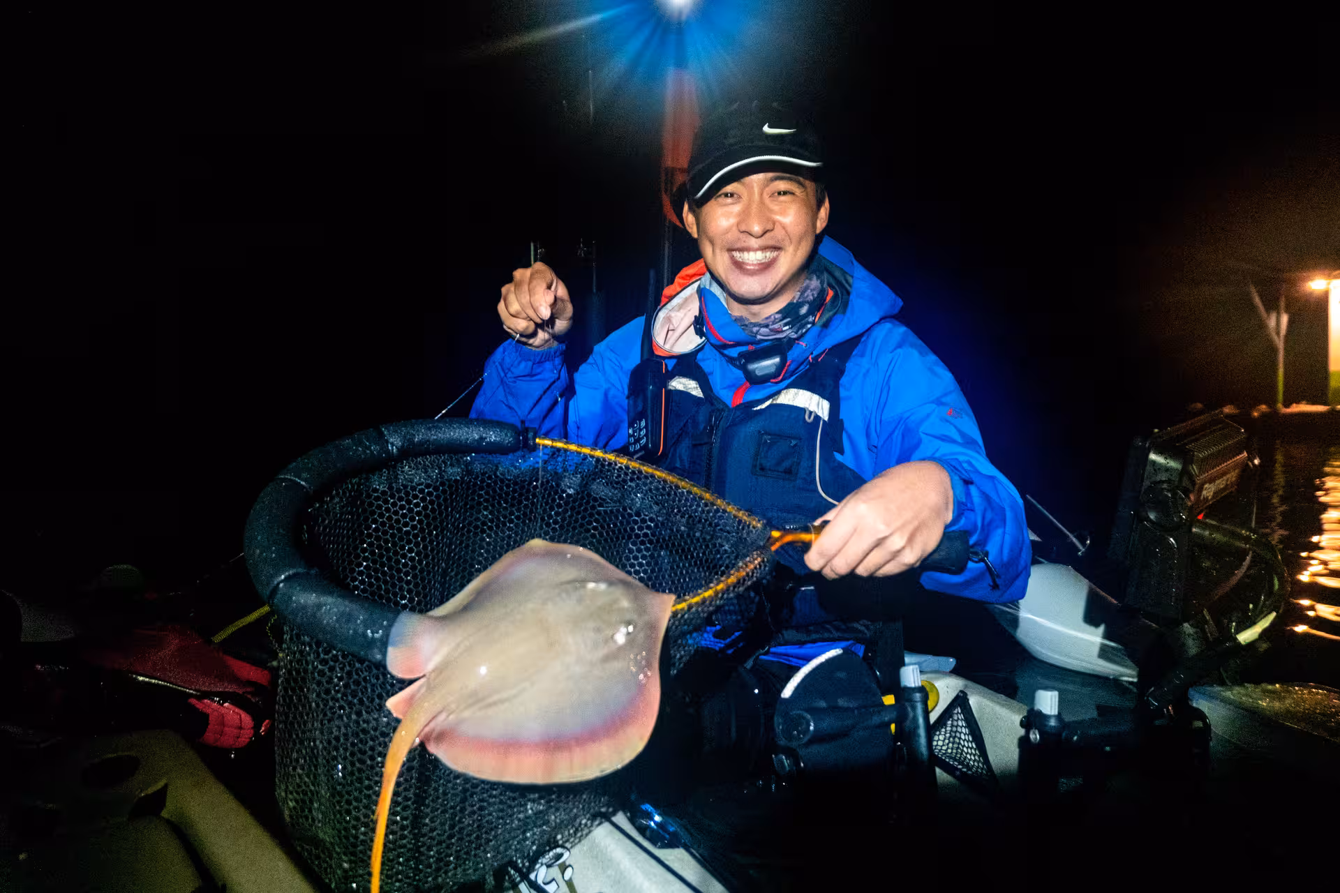 Smiling angler in blue jacket holds a stingray in a net during night kayak fishing at Pulau Ubin.