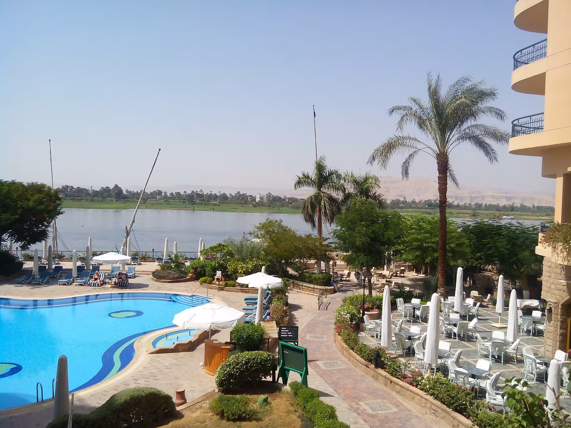 Scenic Nile river view with elegant hotel pool and dining area, framed by palm trees and vibrant gardens.