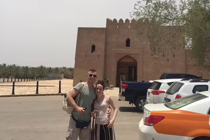 Visitors posing outside the iconic Nizwa Fort, surrounded by palm trees and parked vehicles on a sunny day.