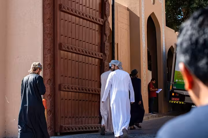 Visitors in traditional dress walk through the grand wooden gate of historic Nizwa Fort, a highlight of the Nizwa by Night sightseeing tour