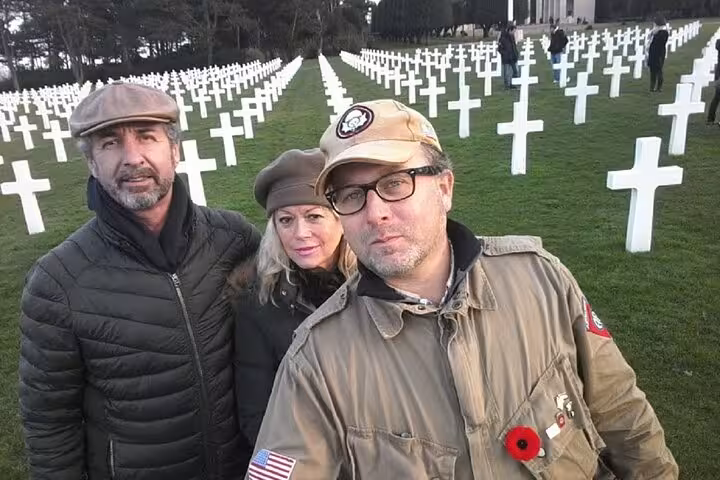 Three visitors posing at Normandy American Cemetery with rows of white crosses behind them