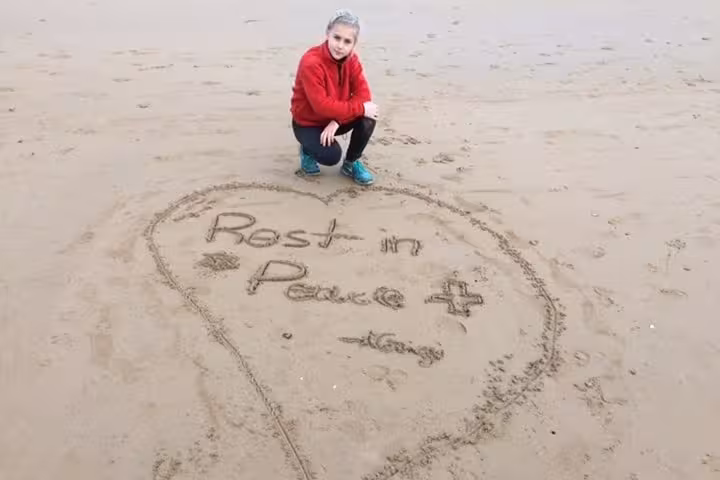Person kneeling beside heart drawn in sand at Normandy beach with Rest in Peace message