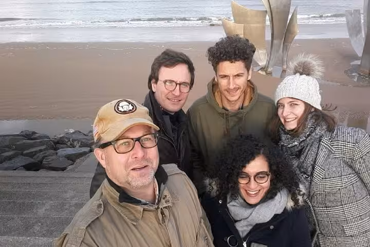 A group of five tourists smiling at a Normandy D-Day beach memorial sculpture during a private tour from Le Havre.
