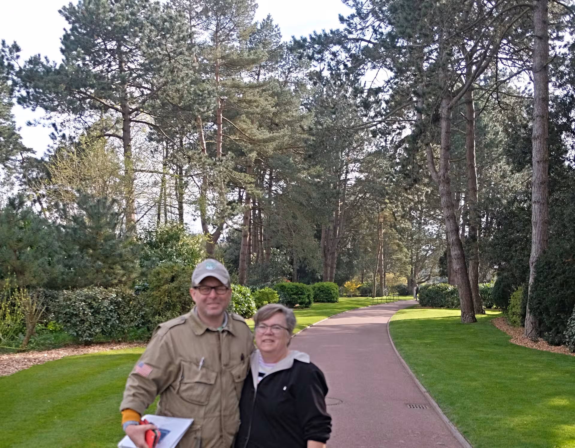 Two tourists posing on a tree-lined path at Normandy D-Day memorial grounds