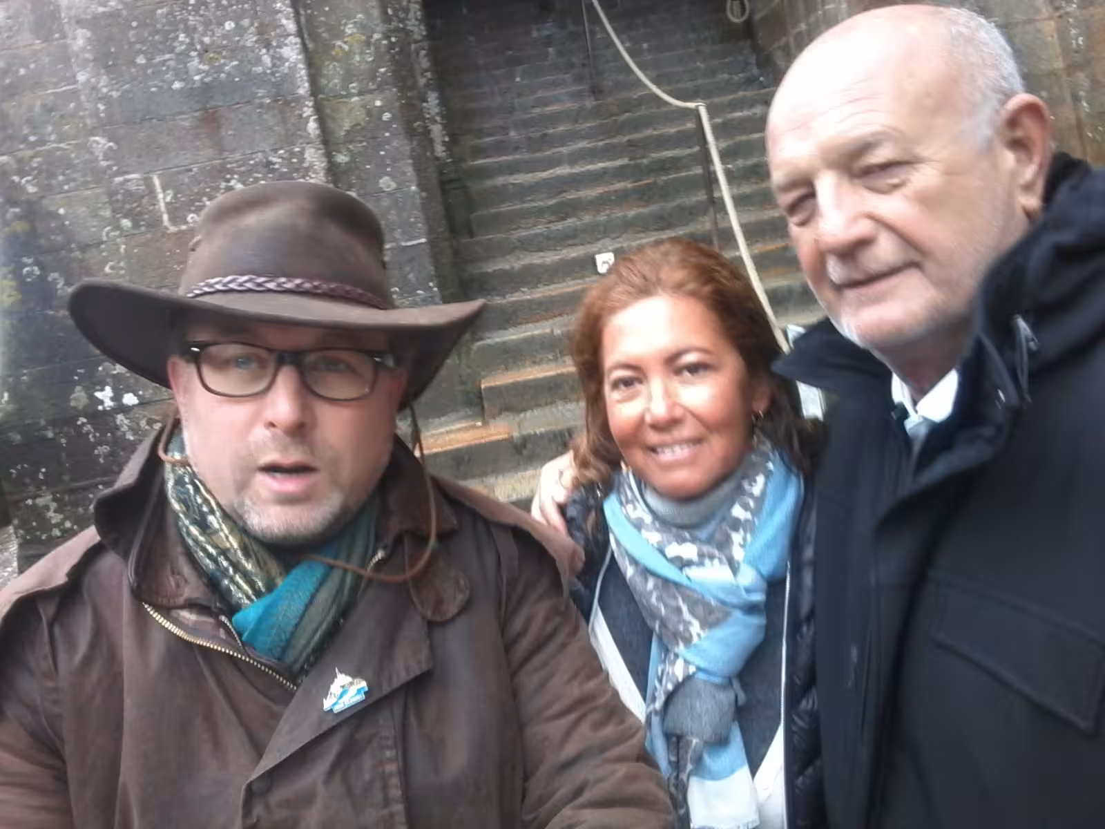 Tour guide with hat and glasses posing with two visitors at historic Normandy stone monument