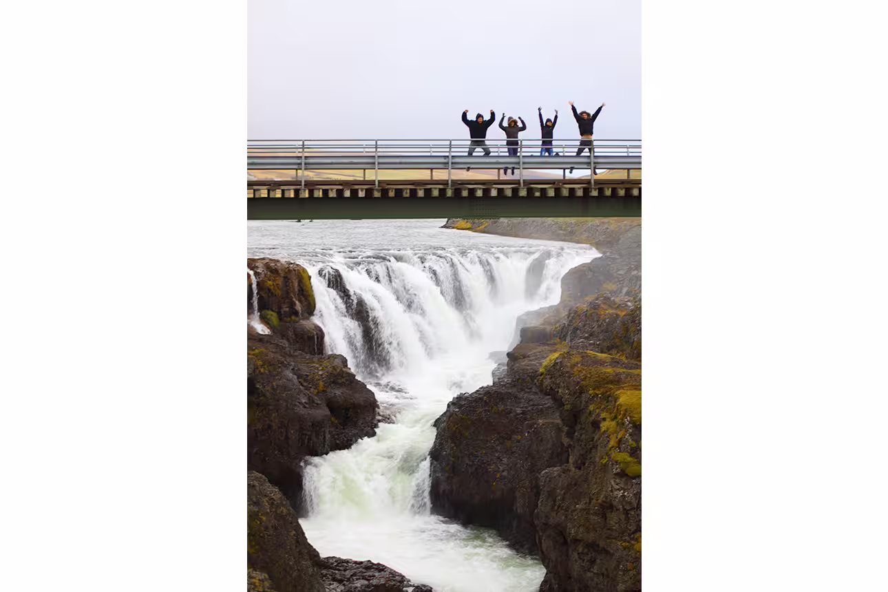 Four tourists jumping on a bridge over a dramatic waterfall and rocky gorge in North Iceland