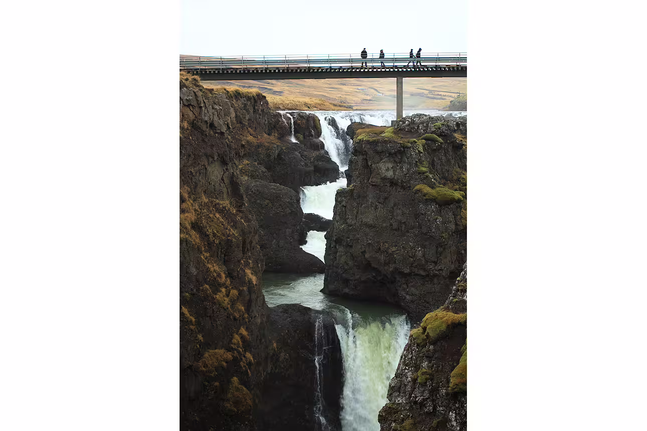 Waterfall cascading through rocky gorge with bridge and tourists overlooking in North Iceland