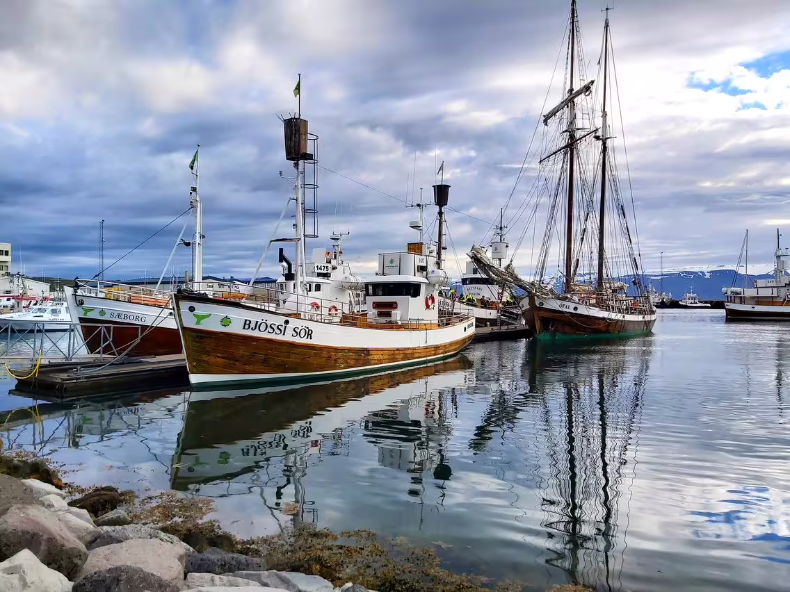 Historic wooden boats docked at a calm harbor in North Iceland under dramatic cloudy skies