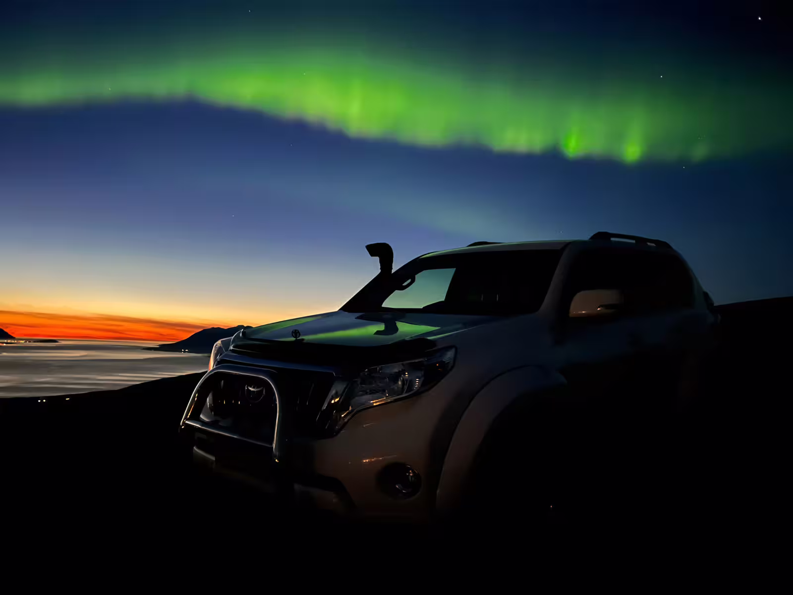 Toyota Land Cruiser SUV parked under vivid green northern lights with sunset glow over calm Arctic waters