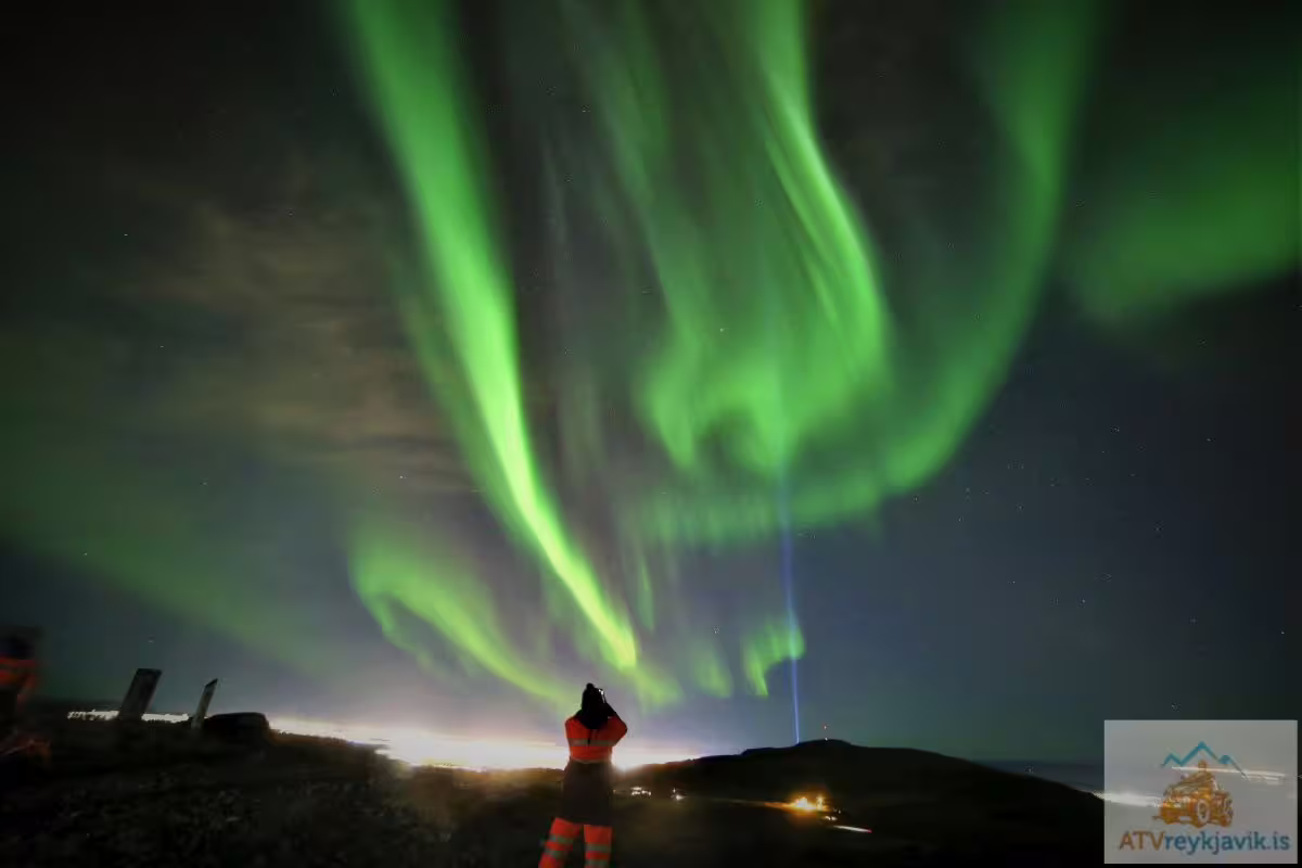 Person in red jacket watching vibrant green northern lights aurora borealis swirling over Reykjavik night sky