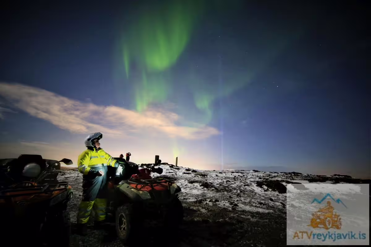 ATV rider in yellow gear watching northern lights aurora borealis over snowy Reykjavik landscape at night