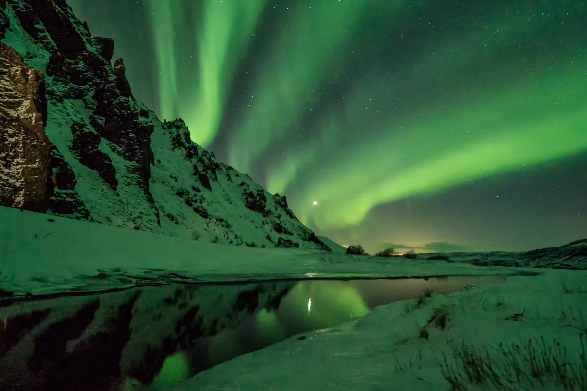 Dramatic green auroras dancing over snowy cliffs and a serene river, ideal for a Northern Lights 4x4 tour.