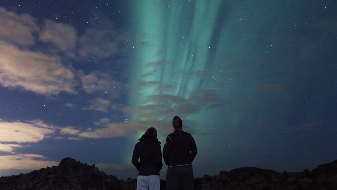 Two guests stand in awe watching vibrant green northern lights aurora borealis dance across the starry night sky on a guided hunt tour