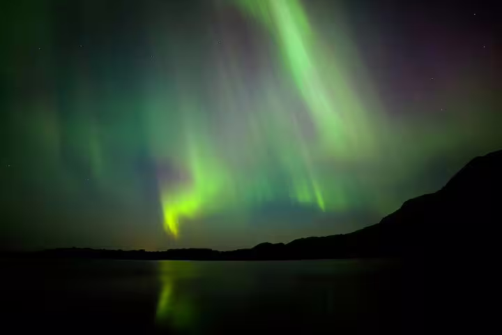 Vibrant green aurora borealis dancing over a calm reflective lake with silhouetted mountains