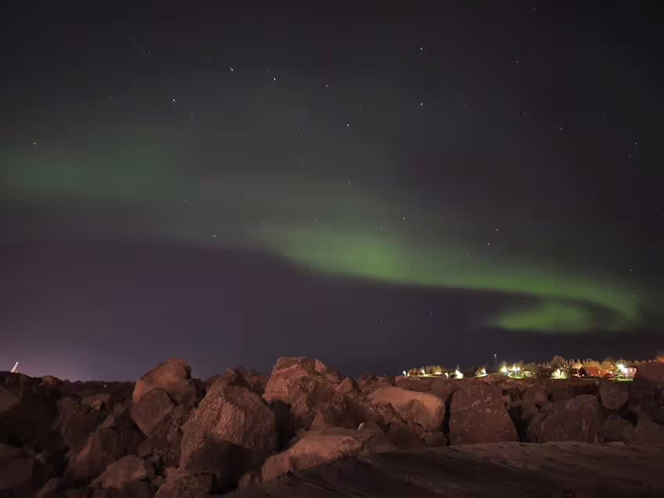 Green aurora borealis dancing over rocky shoreline with distant town lights at night