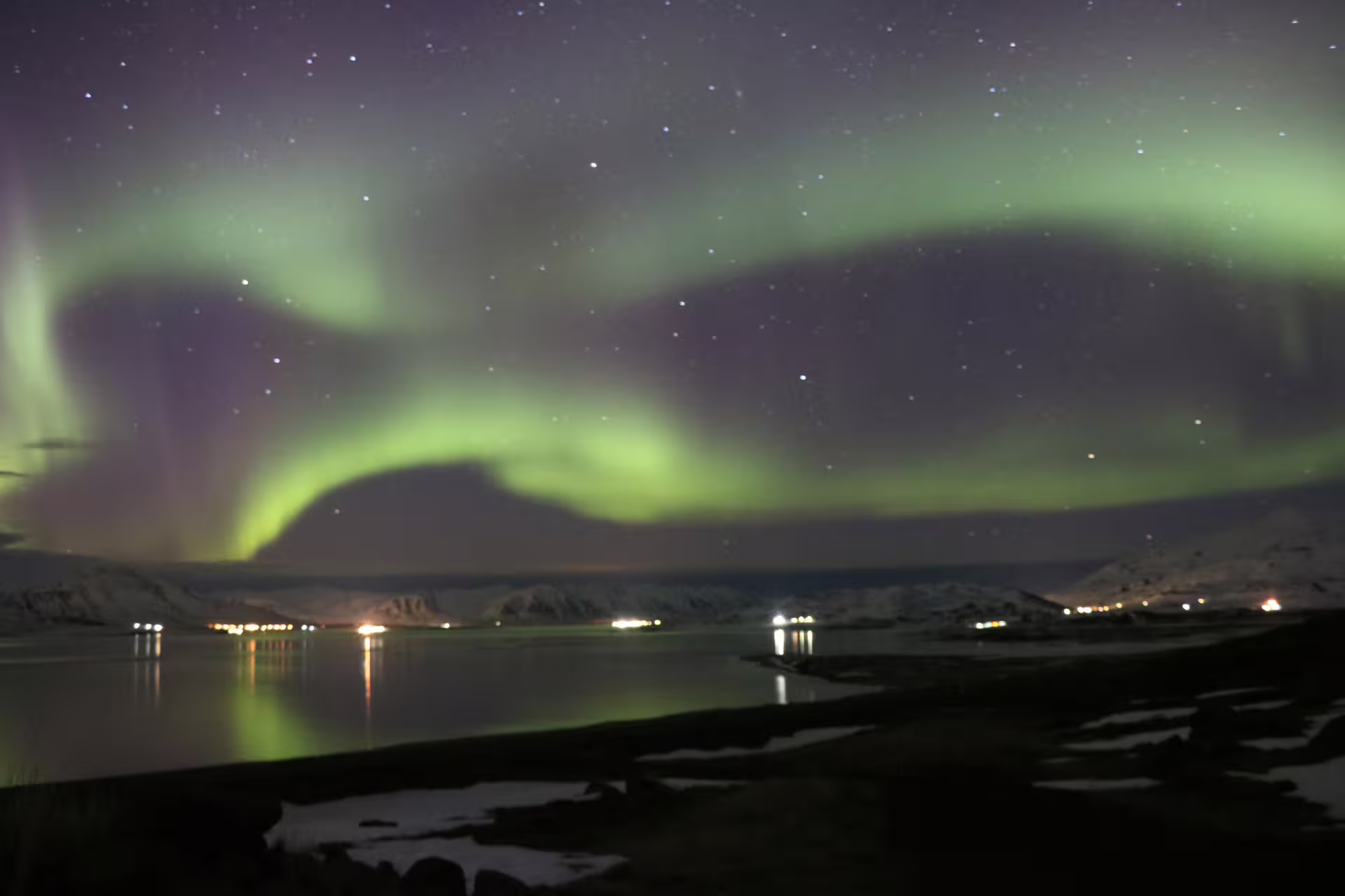 Vibrant green northern lights aurora borealis over a snowy Icelandic fjord with glowing town lights reflected in water