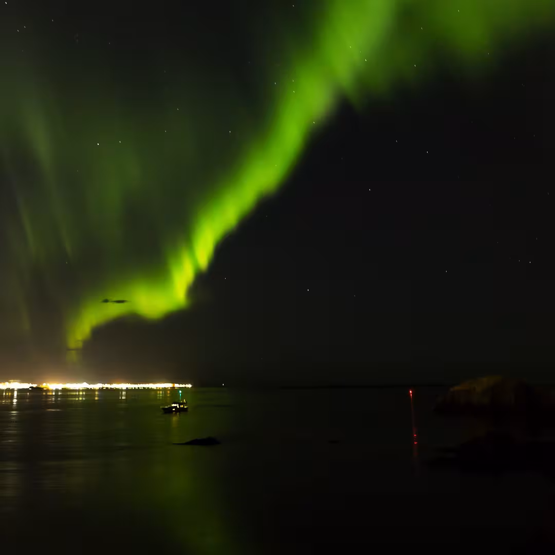 Brilliant green northern lights aurora borealis reflecting over calm Arctic waters with cruise boat and coastal town lights