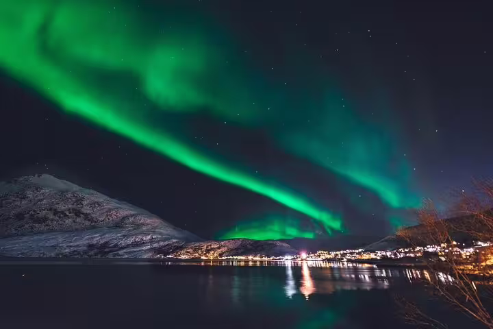Vibrant green Northern Lights dancing over a fjord with snow-capped mountains and city lights reflected in calm water