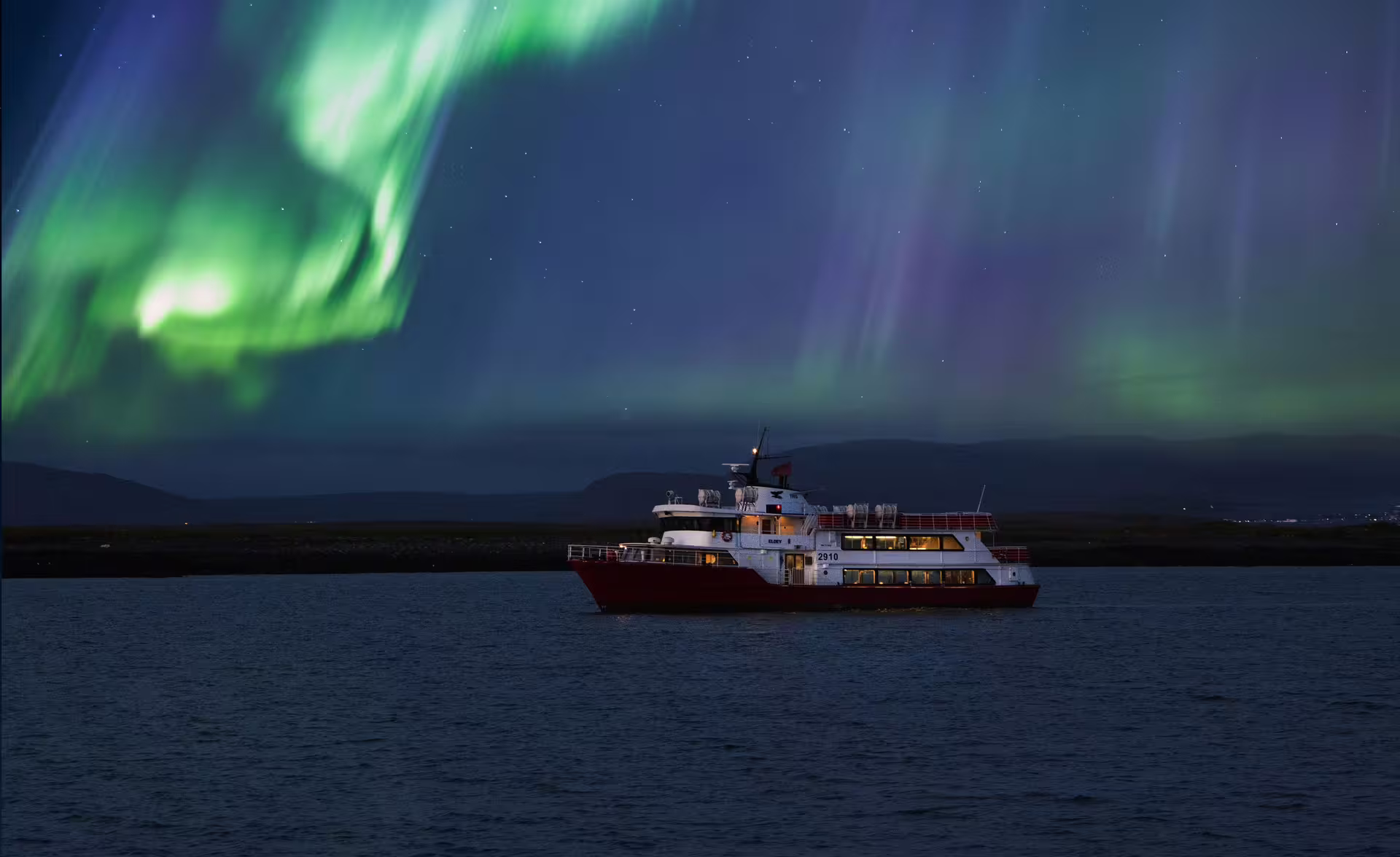 Red and white cruise boat sailing under vivid green and purple northern lights in Reykjavik winter night sky