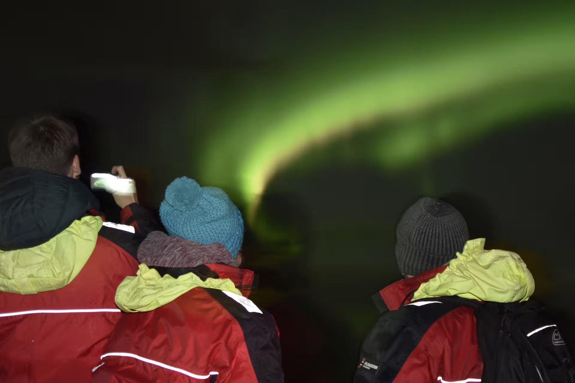 Tourists in red jackets watching vibrant green northern lights aurora borealis from a boat in Reykjavik