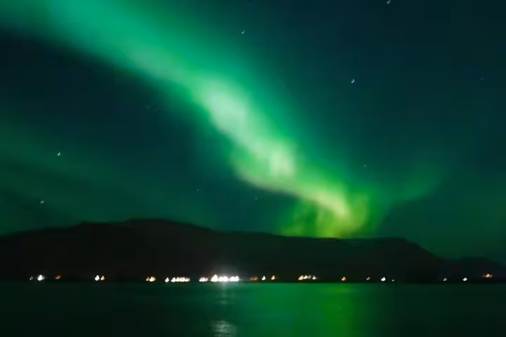 Vibrant green northern lights aurora borealis over a fjord with town lights reflecting on calm water in Iceland