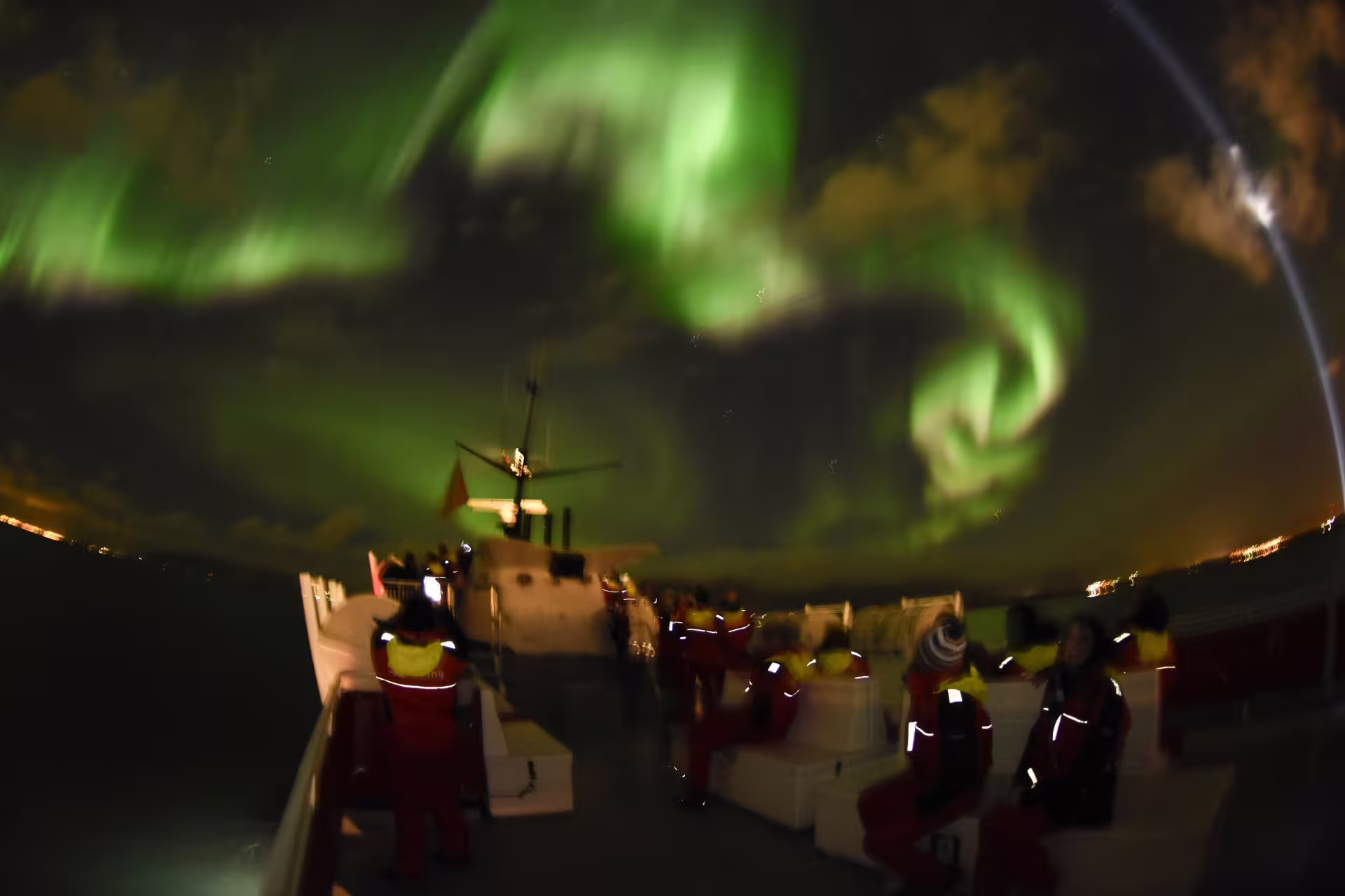 Guests in red survival suits on boat deck watching vivid green northern lights swirl over Reykjavik Bay at night