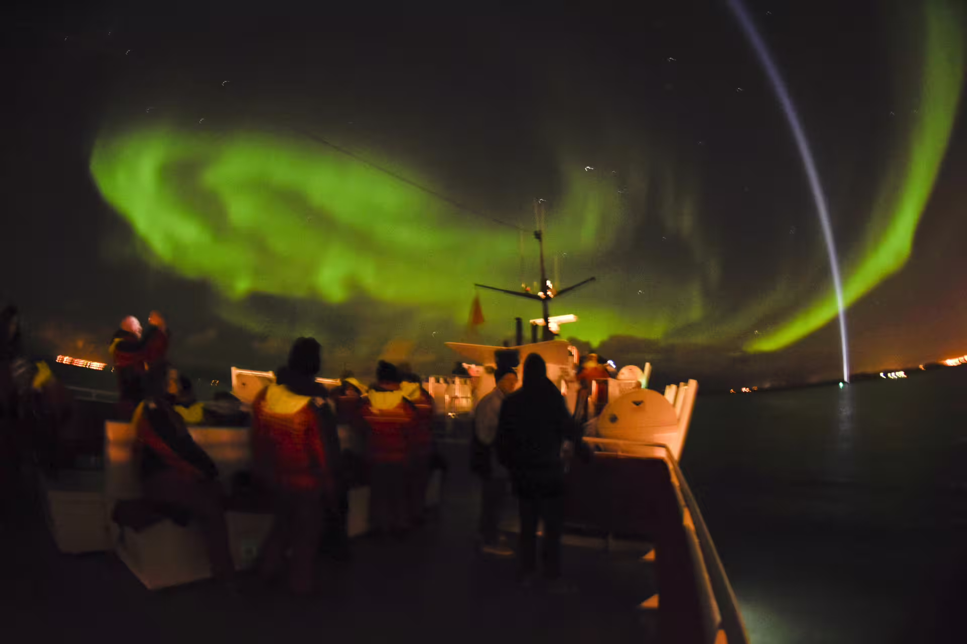 Tourists on boat deck watching vibrant green northern lights swirling over Reykjavik Bay at night