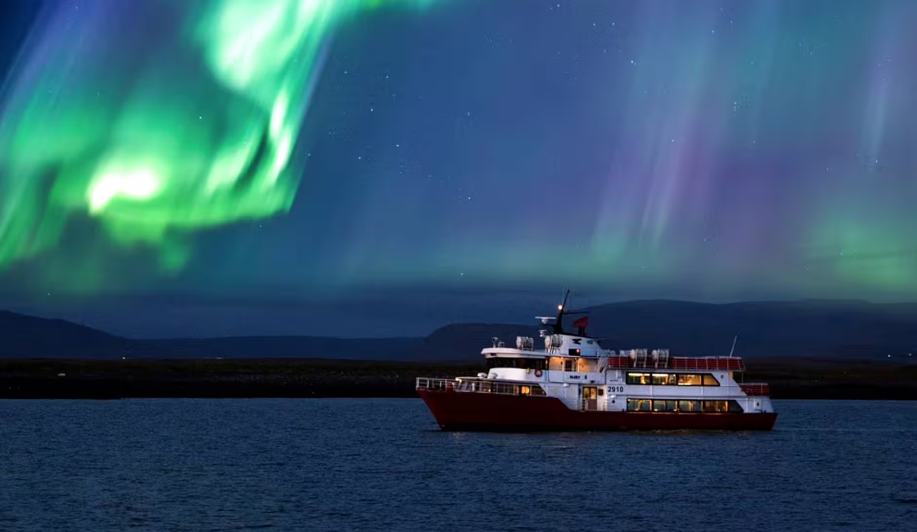 Red and white boat cruising Reykjavik Bay under vibrant green northern lights aurora borealis at night