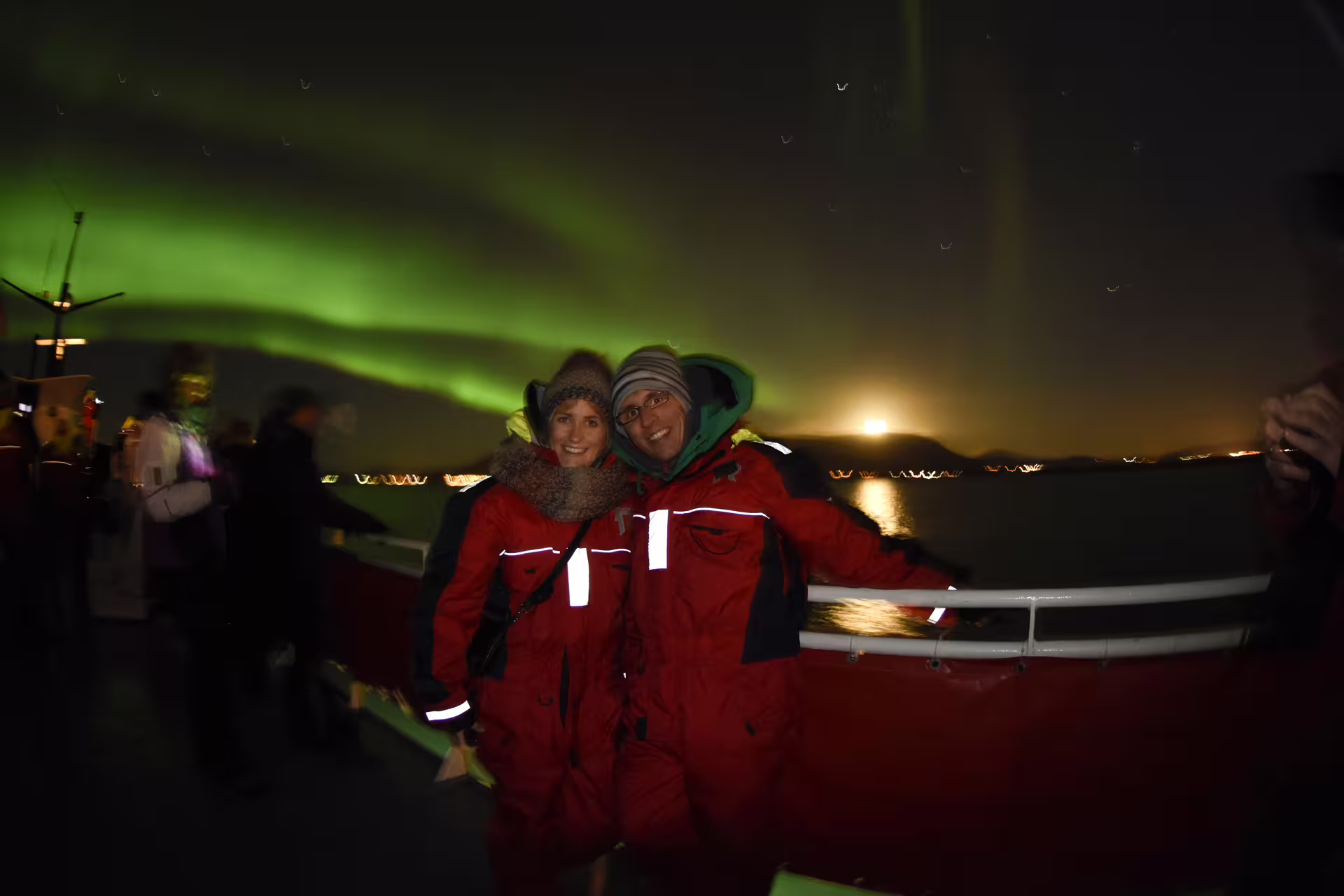 Couple in red survival suits posing on boat with northern lights and moon over Reykjavik Bay at night