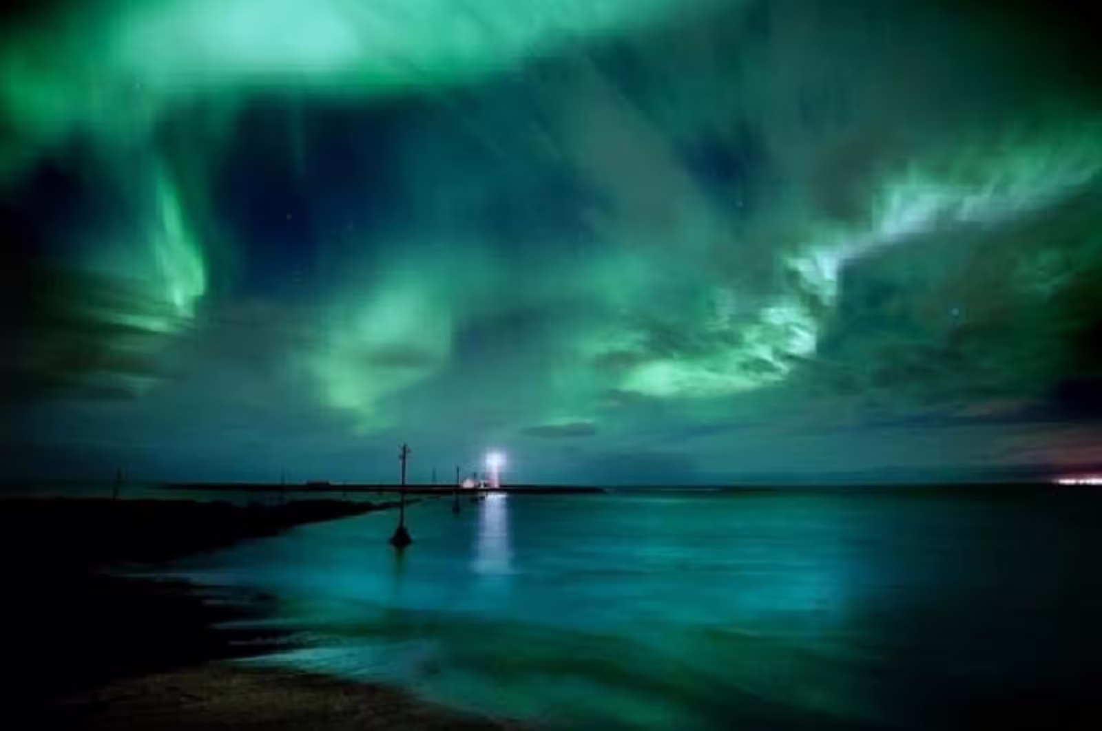 Aurora borealis northern lights glowing green over calm ocean waters with lit pier in Reykjavik Iceland at night