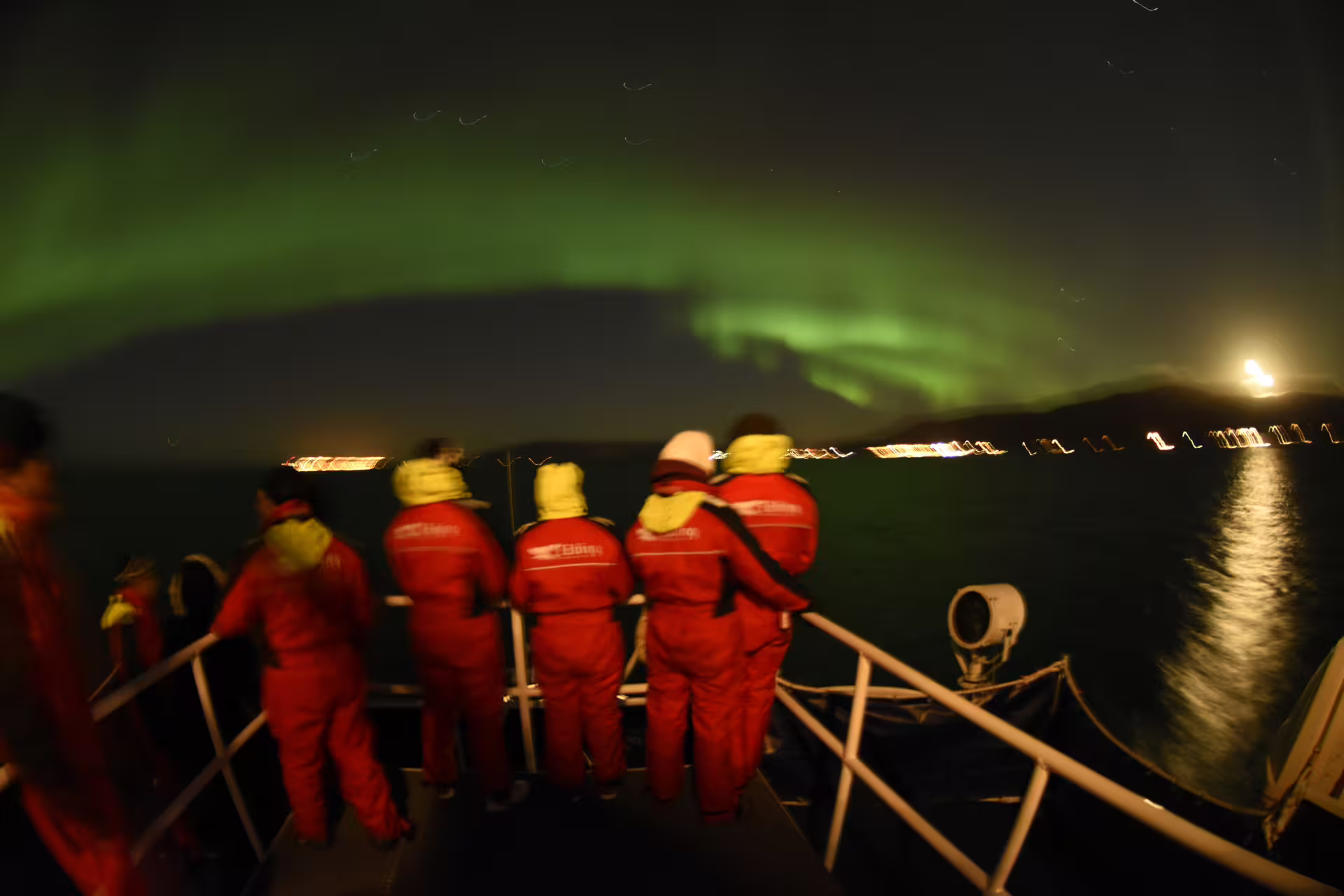 Tourists in red survival suits watching northern lights from a boat deck in Reykjavik at night
