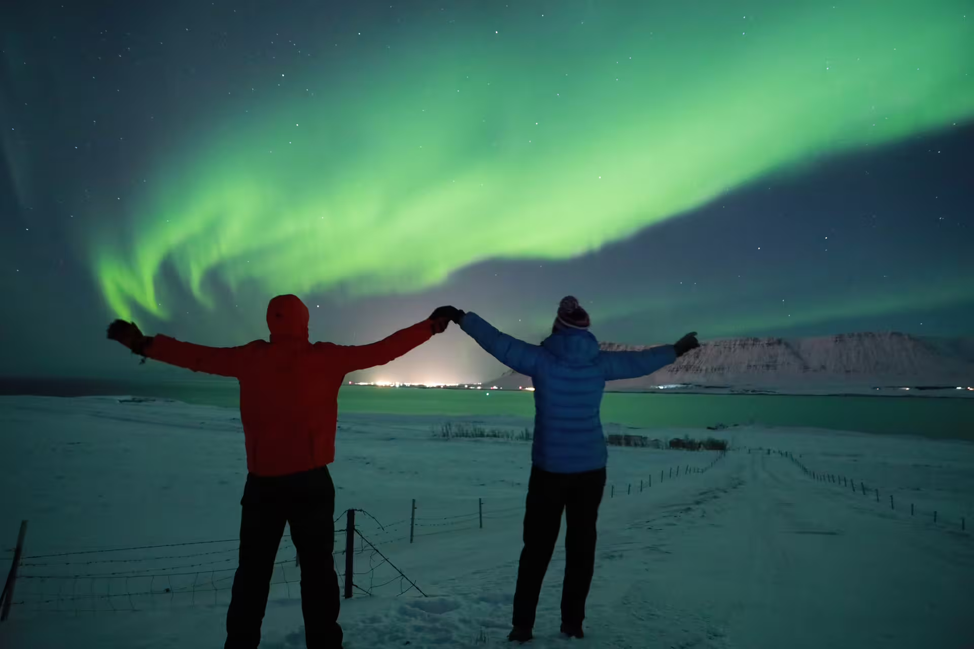 Couple holding hands under vibrant green northern lights over snowy Icelandic landscape at night