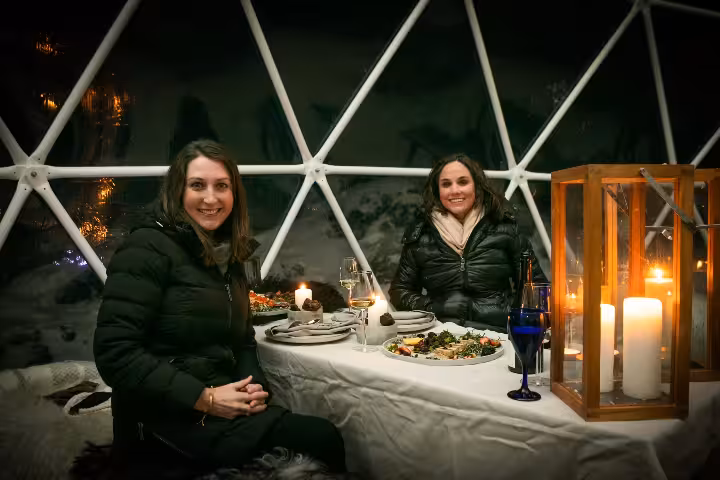 Two women enjoying a candlelit dinner inside a cozy geodesic dome igloo under the northern lights in Iceland