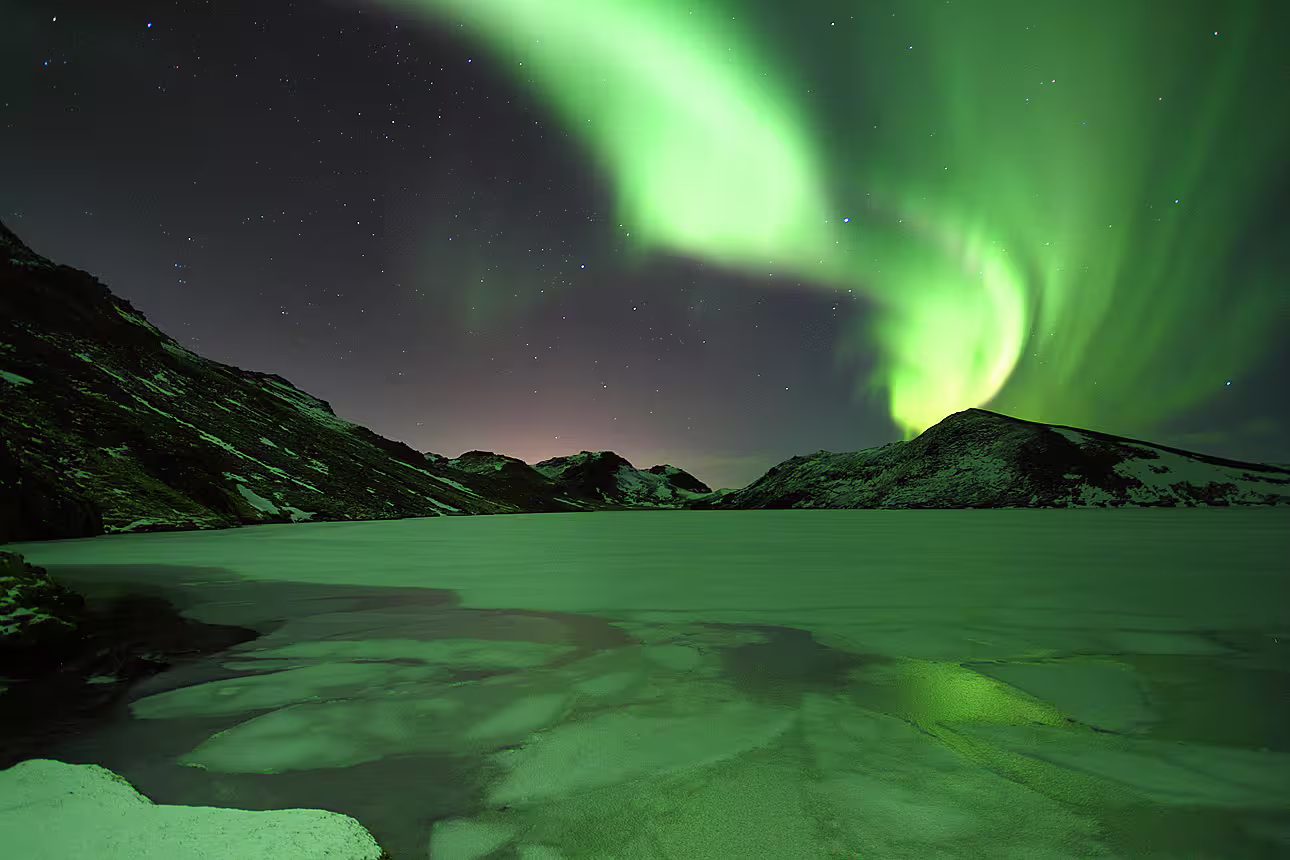 Vivid green northern lights swirling over a frozen lake surrounded by snow-covered mountains in Iceland