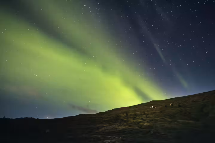 Vivid green northern lights aurora borealis sweeping over a dark hillside with a small lit cabin in Iceland