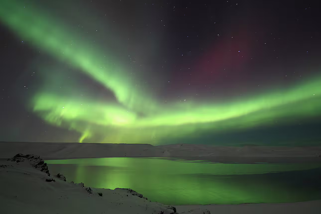 Vivid green and red northern lights reflecting over a snow-covered lake in Iceland at night