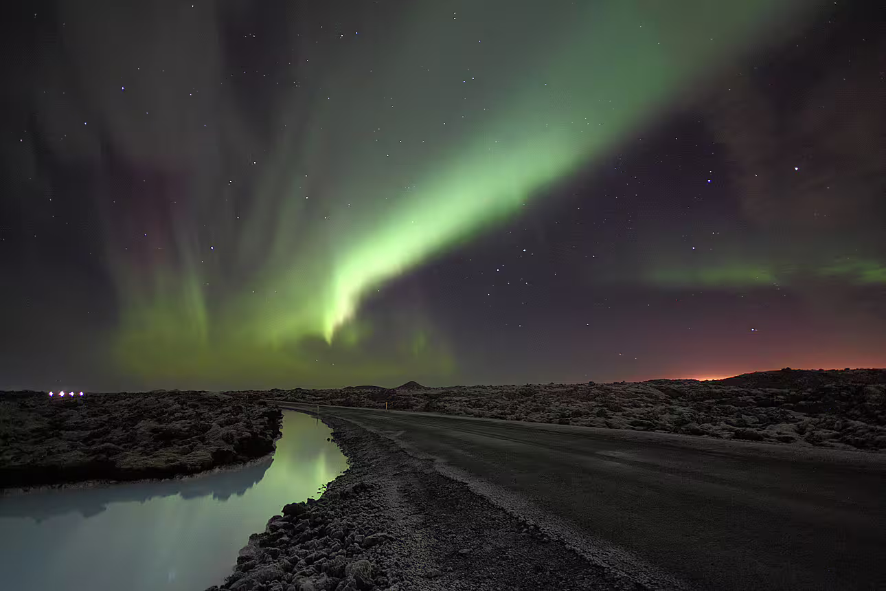 Aurora borealis dancing over a road and reflective canal in Iceland's volcanic landscape at night