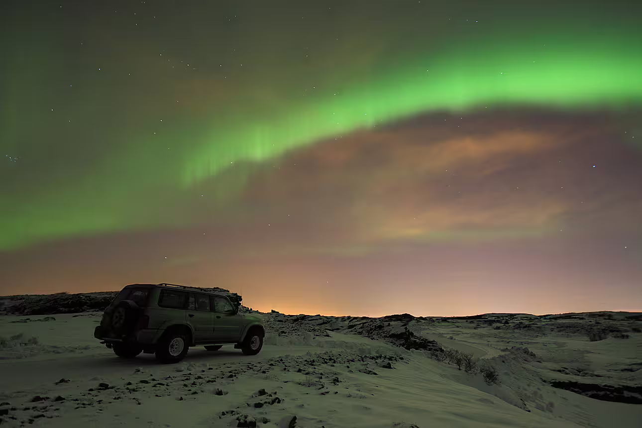 4WD SUV parked on snowy landscape under vivid green northern lights in Iceland night sky